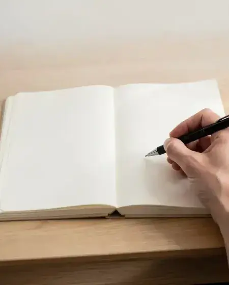 Hands holding a pen over a blank open notebook on a wooden desk in soft light