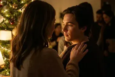 A mother gently touching her adult child's shoulder by a warm Christmas tree, symbolizing the shelter of home and the enduring strength of love.