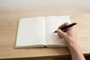 Hands holding a pen over a blank open notebook on a wooden desk in soft light